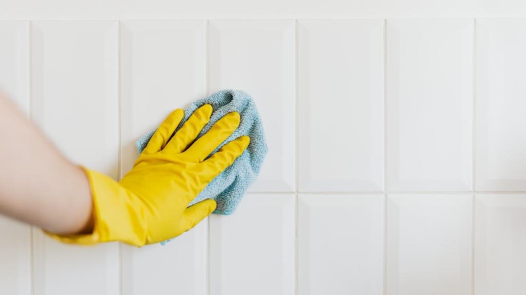 Gloved hand wiping a white tile wall with a microfiber cloth
